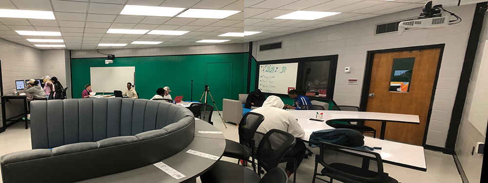 Panoramic view of a modern study room with students working at various stations. The room features a large, semi-circular seating area in the center, students using computers on the left, and others working on laptops and whiteboards on the right. The walls are painted green and gray, with ample overhead lighting.